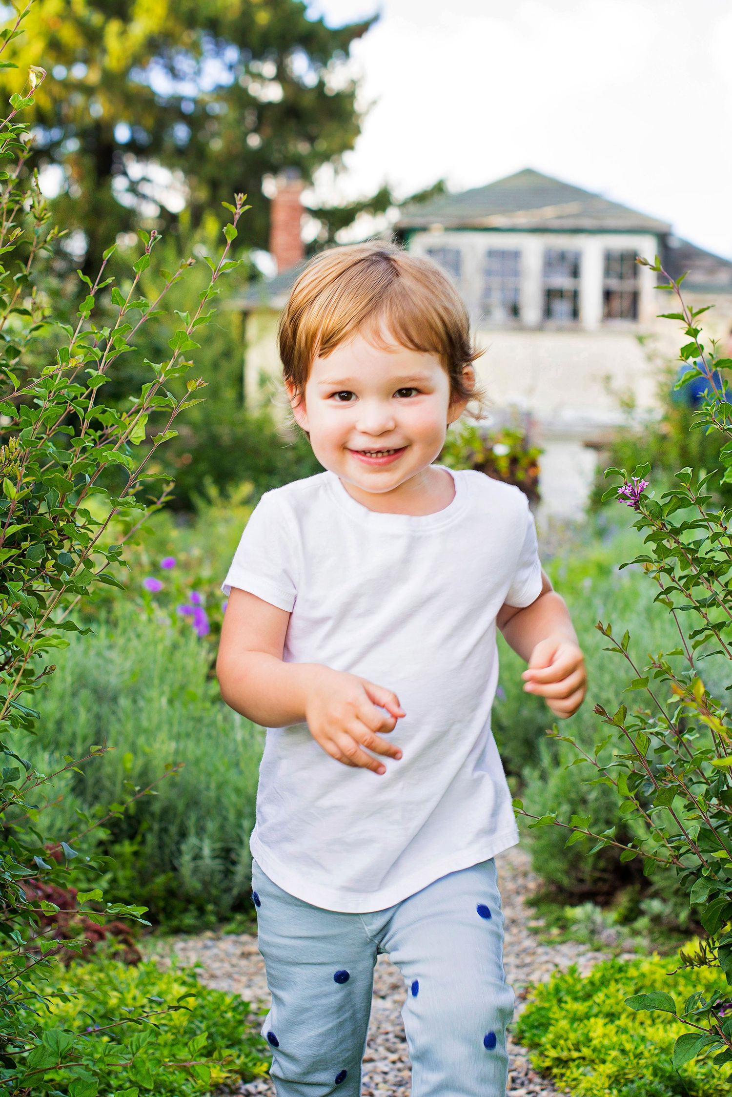 Happy toddler in white shirt running through a lush green garden path on a sunny day.