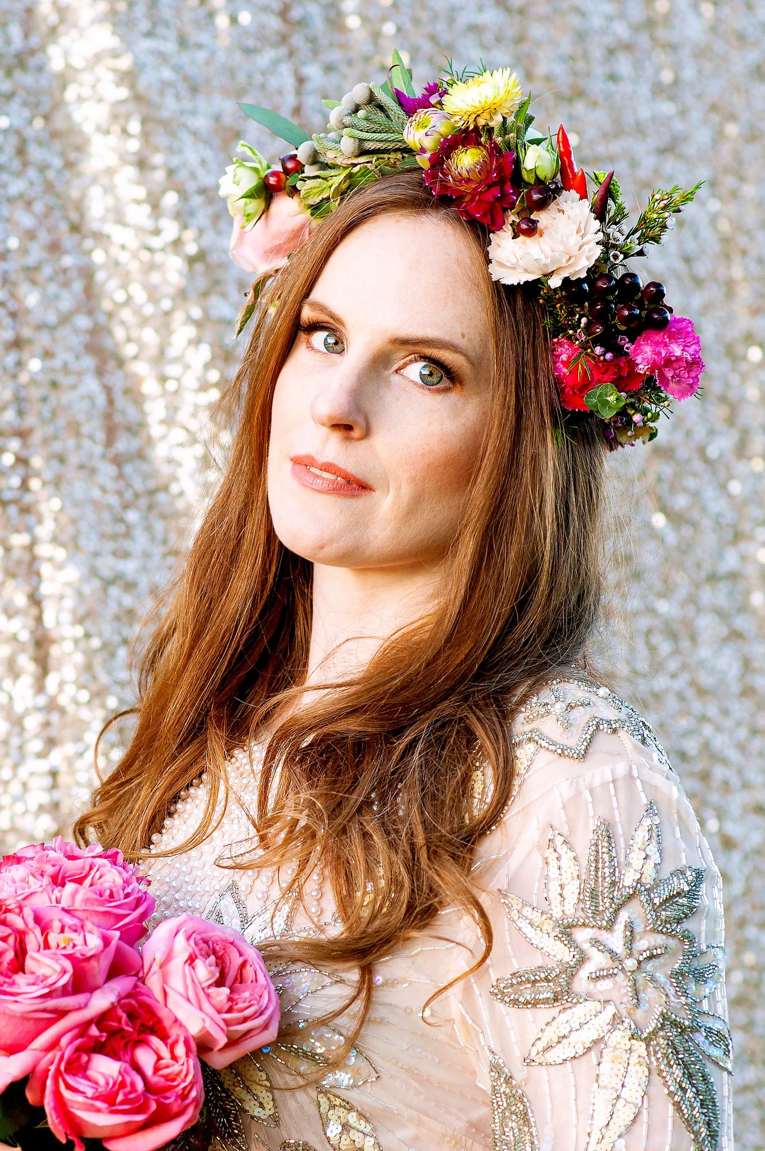 Beautiful woman with vibrant floral crown holding pink roses bouquet against silver backdrop.