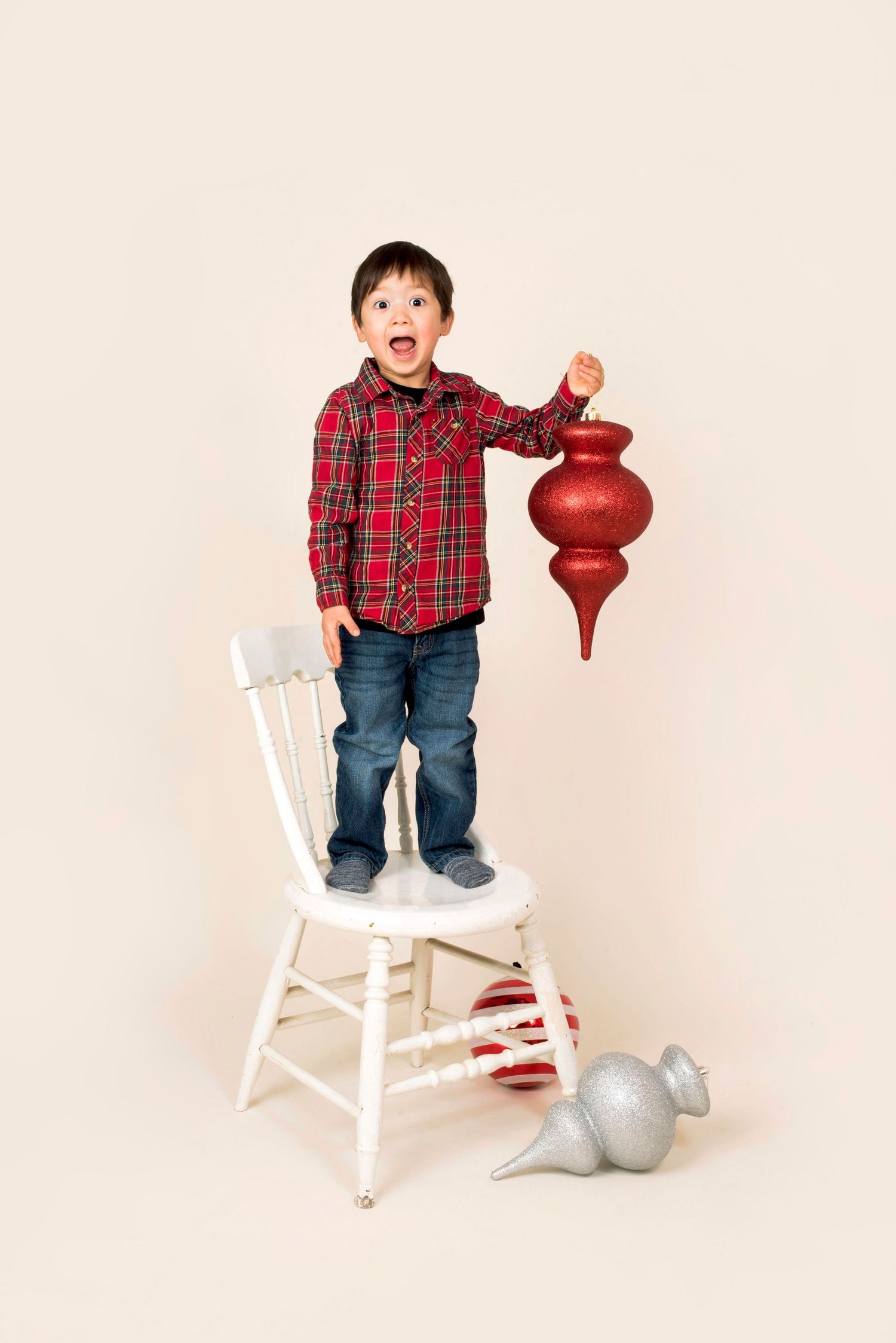 Excited young boy standing on white chair holding large red Christmas ornament.