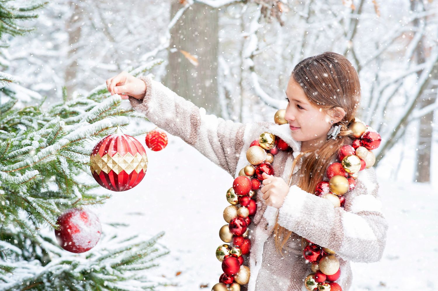 Young girl decorating snowy Christmas tree with red and gold ornaments.