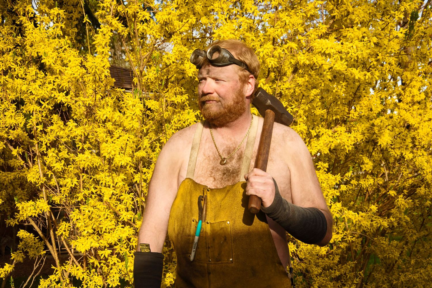 Shirtless bearded man wearing a leather apron and headlamp holds a hammer before bright yellow flowers.