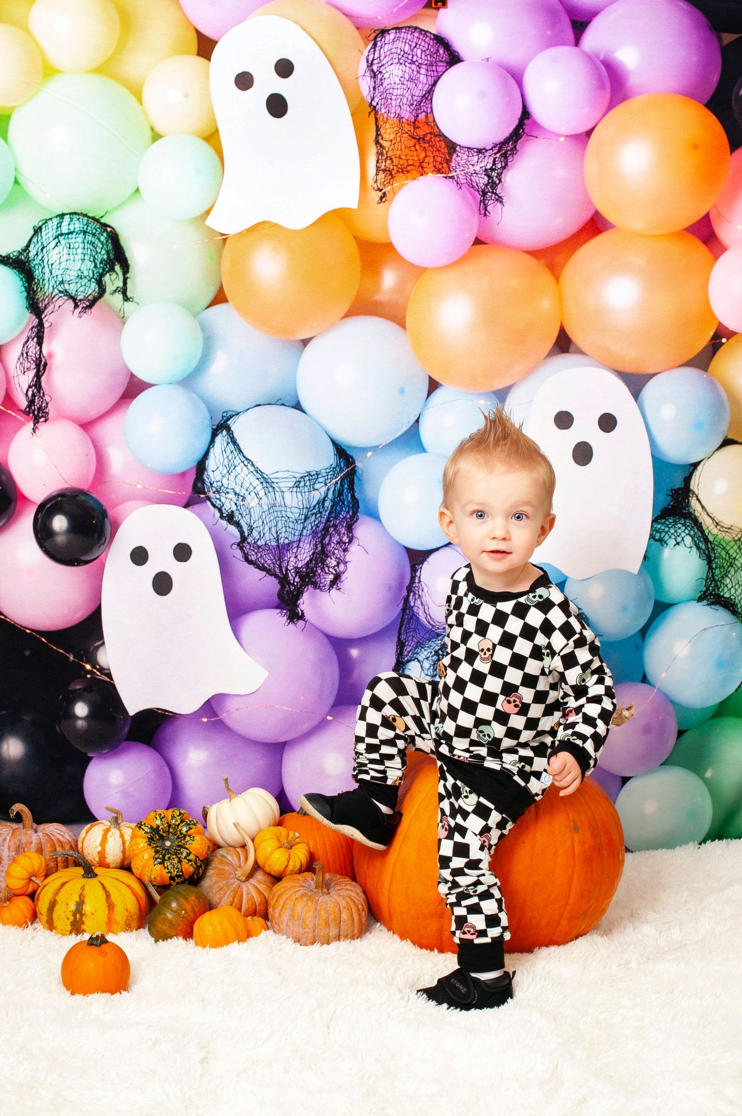 Toddler in checkered outfit sitting on pumpkin with colorful Halloween balloon backdrop.