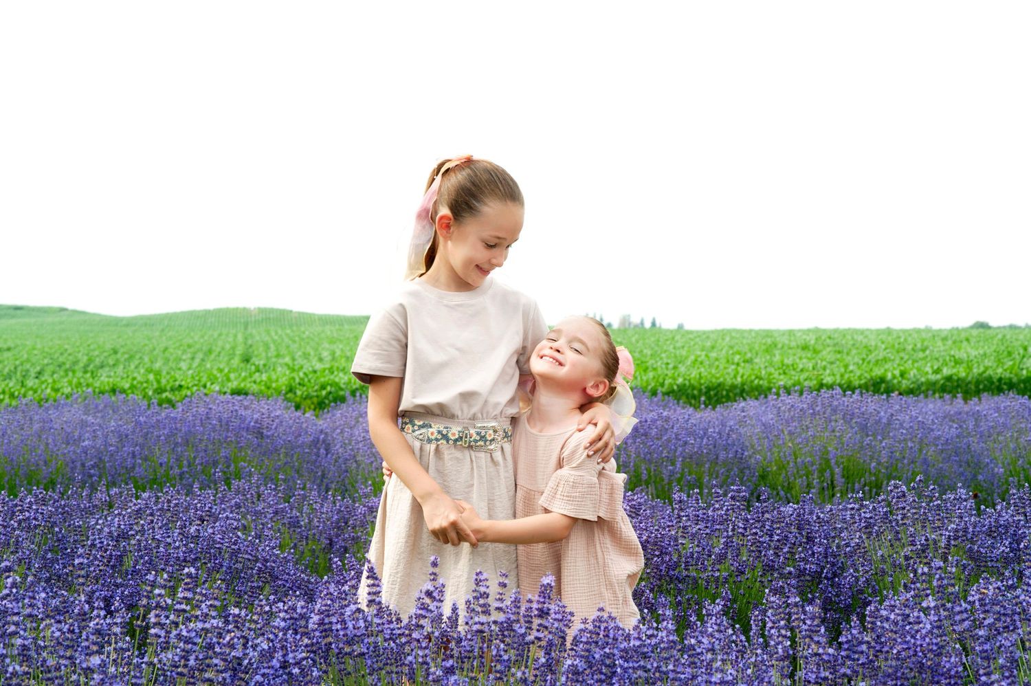 Two young girls in matching beige dresses embrace joyfully in a blooming purple lavender field.