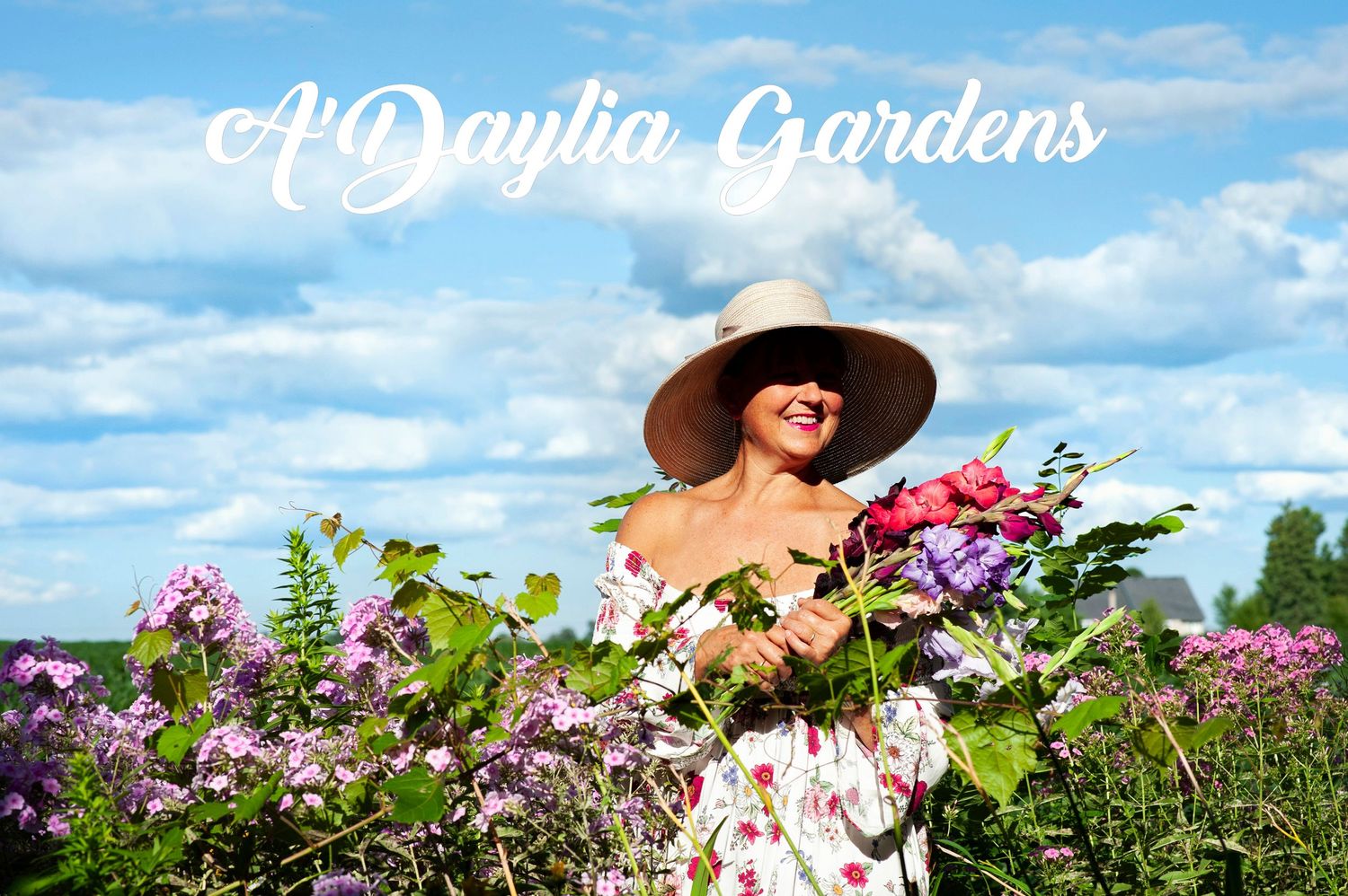 Smiling woman in white floral dress and sun hat surrounded by pink and purple flowers at A Daylia Gardens.