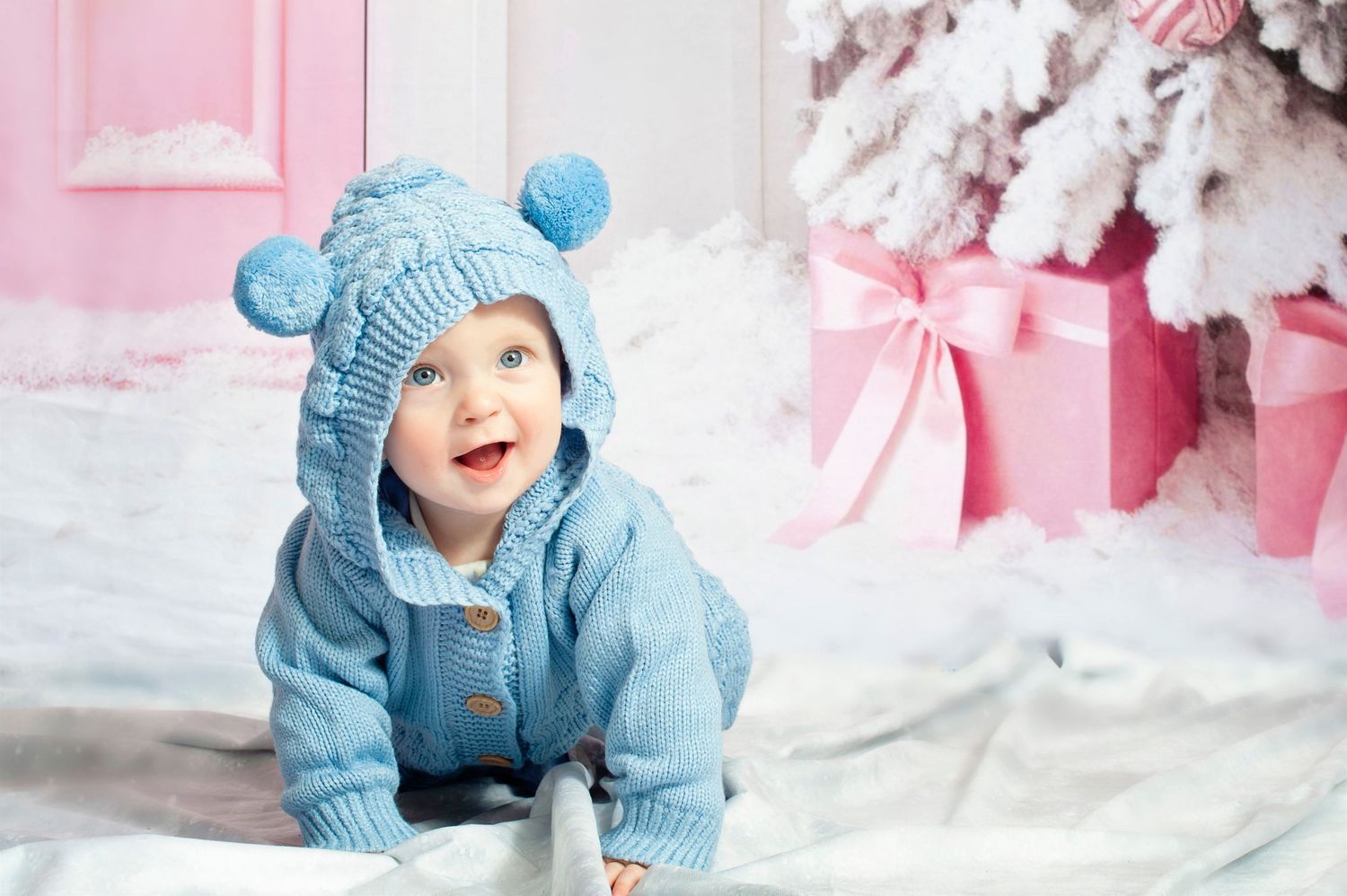 Happy baby in blue knit bear hat crawling near Christmas tree with pink gifts.