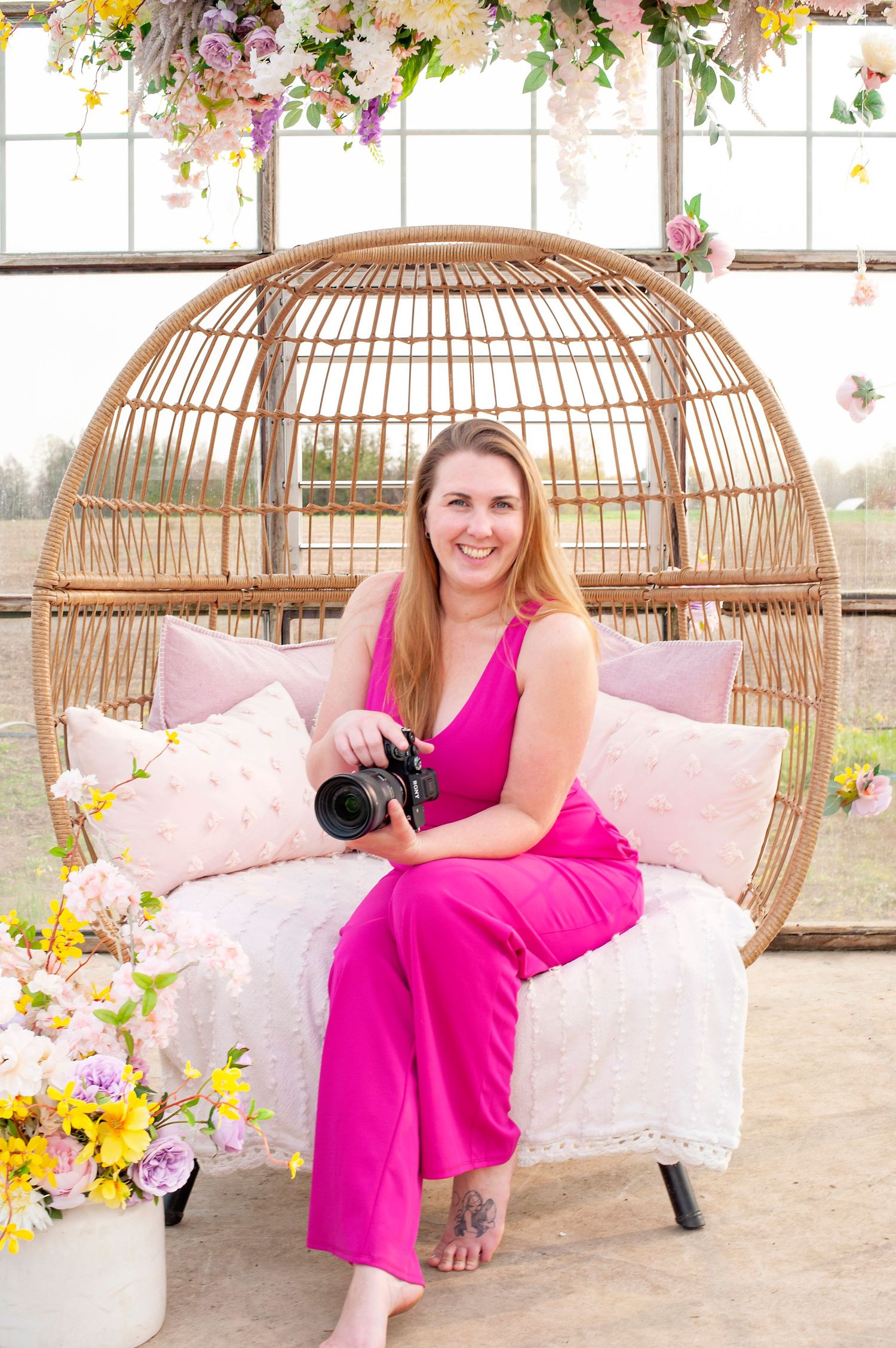 Smiling female photographer in pink jumpsuit holding camera sits in rattan egg chair with floral decor in greenhouse.
