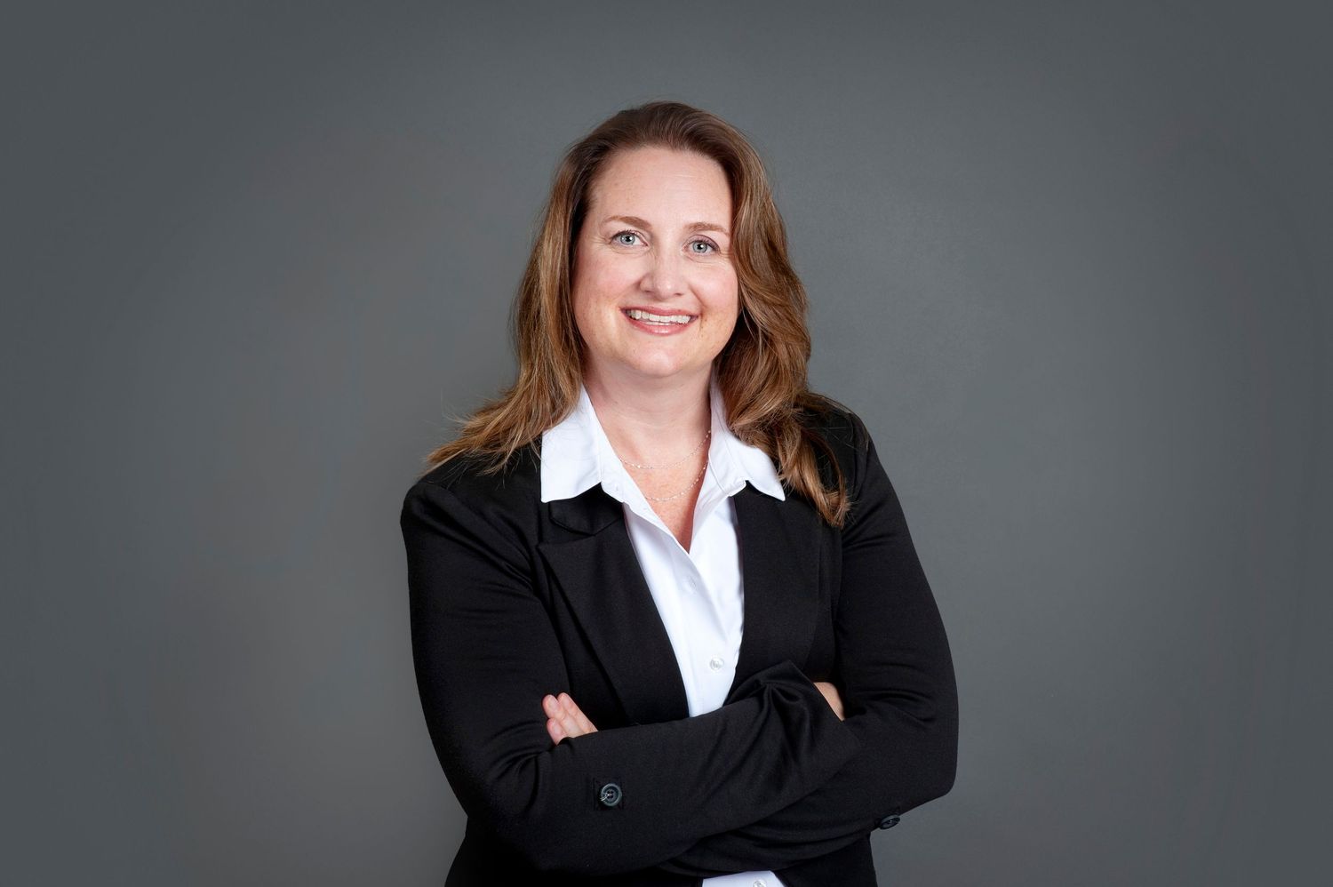 Professional woman in black blazer poses with arms crossed against gray background.