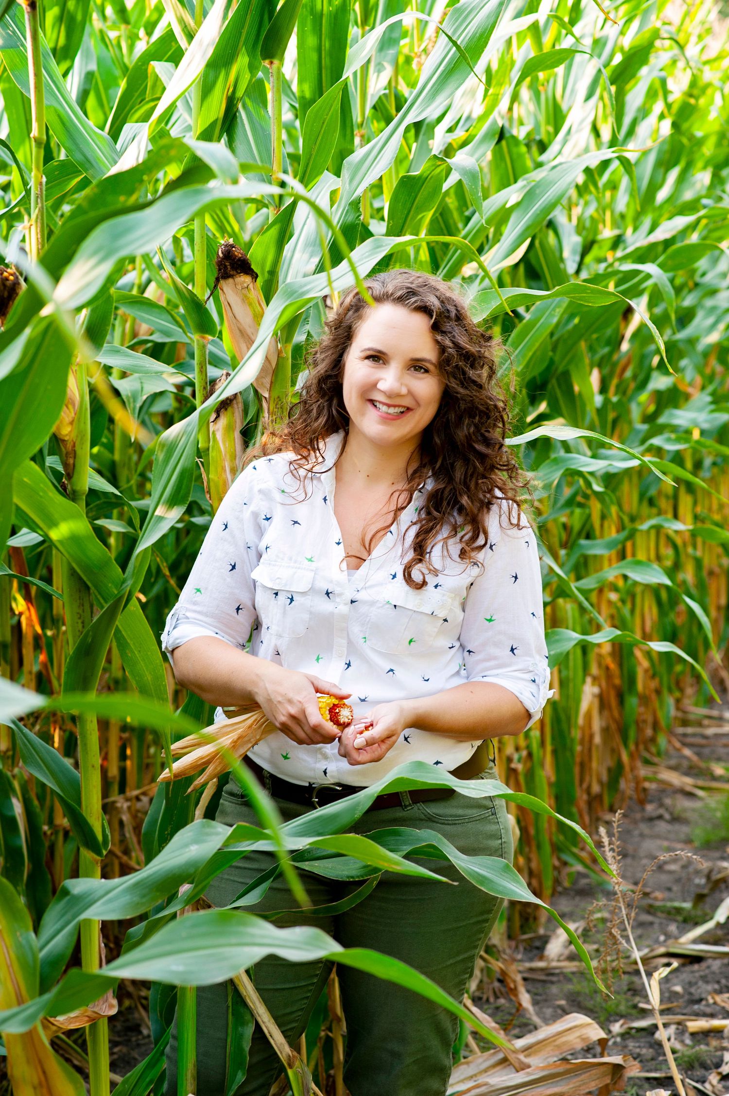 Smiling woman with curly hair holding corn in a lush green cornfield.