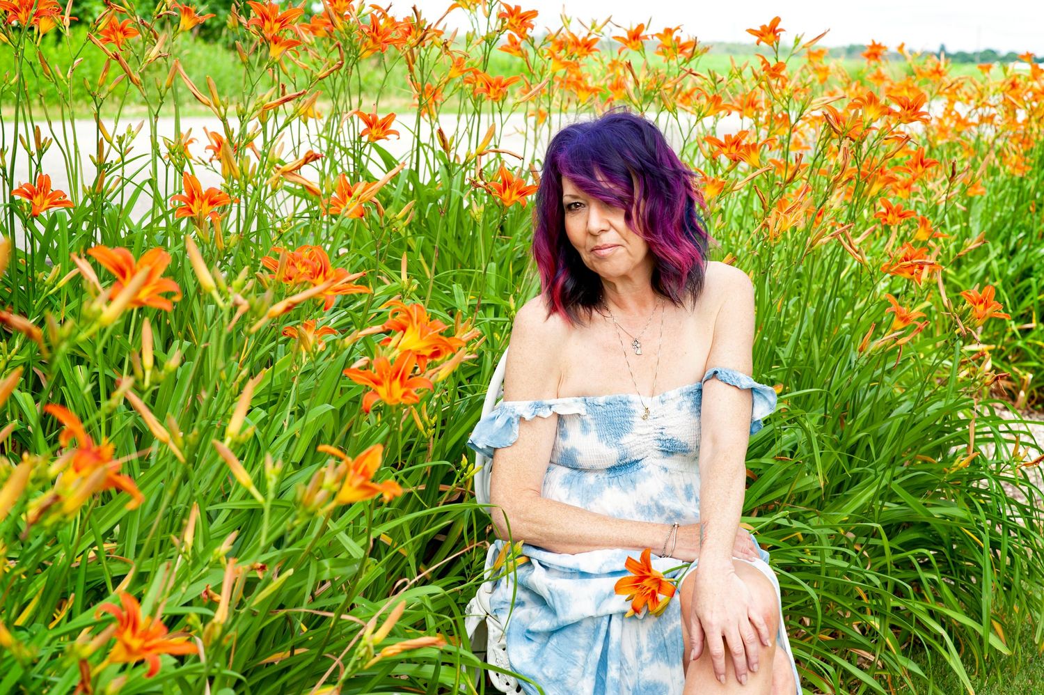 Woman with purple hair in denim dress posing near orange lily flowers.