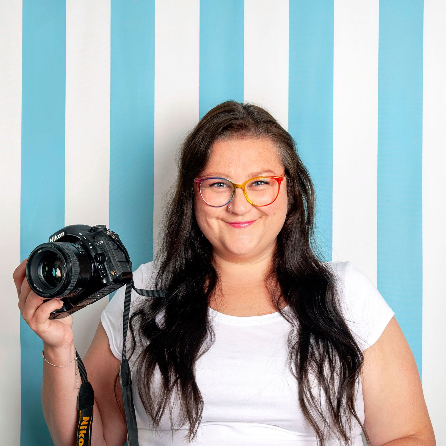 Smiling brunette woman holds a Nikon camera in front of a blue and white striped background.