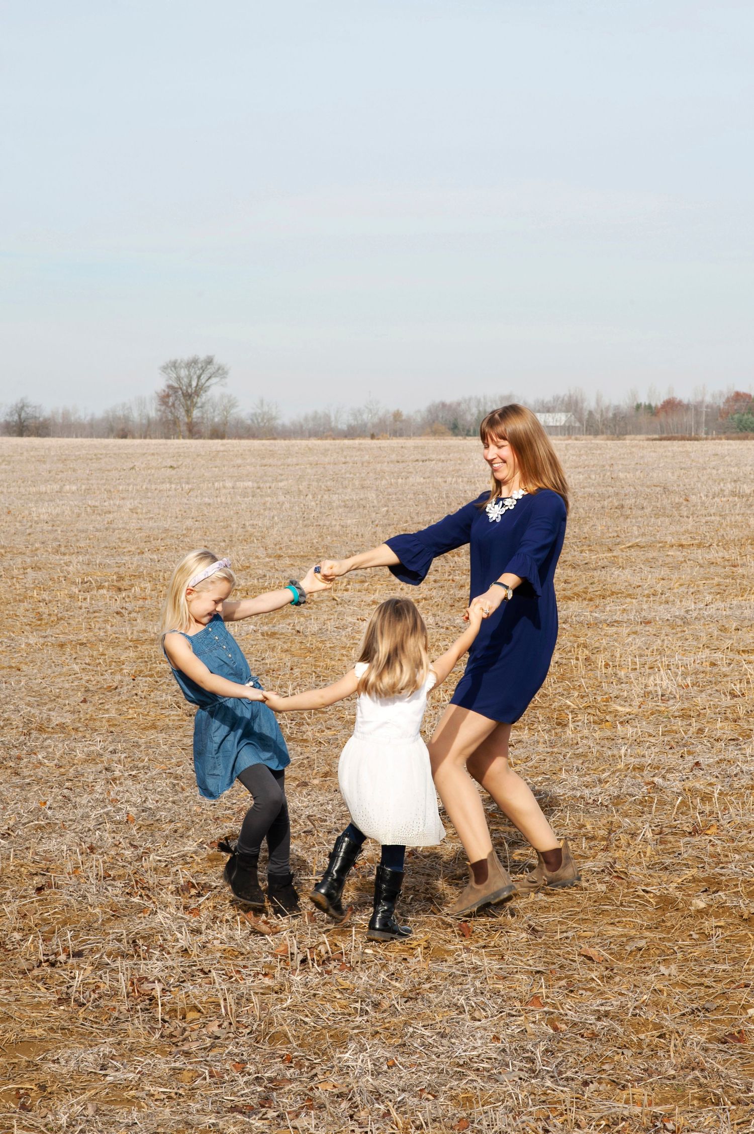 Woman in blue dress spinning and dancing with two young blonde girls in an open autumn field.