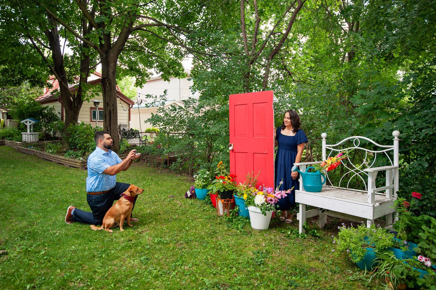 Man proposing to woman near a red door with their dog and colorful flowers in garden.