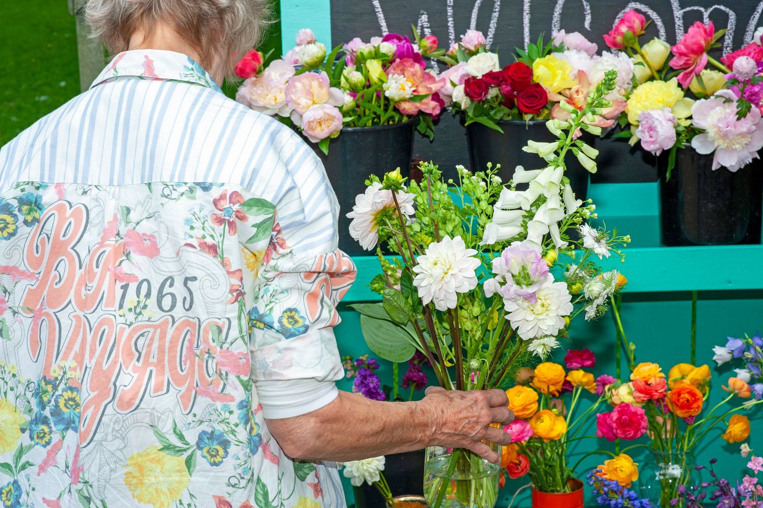 Elderly woman in floral shirt selecting flowers from a colorful outdoor flower market display stand.