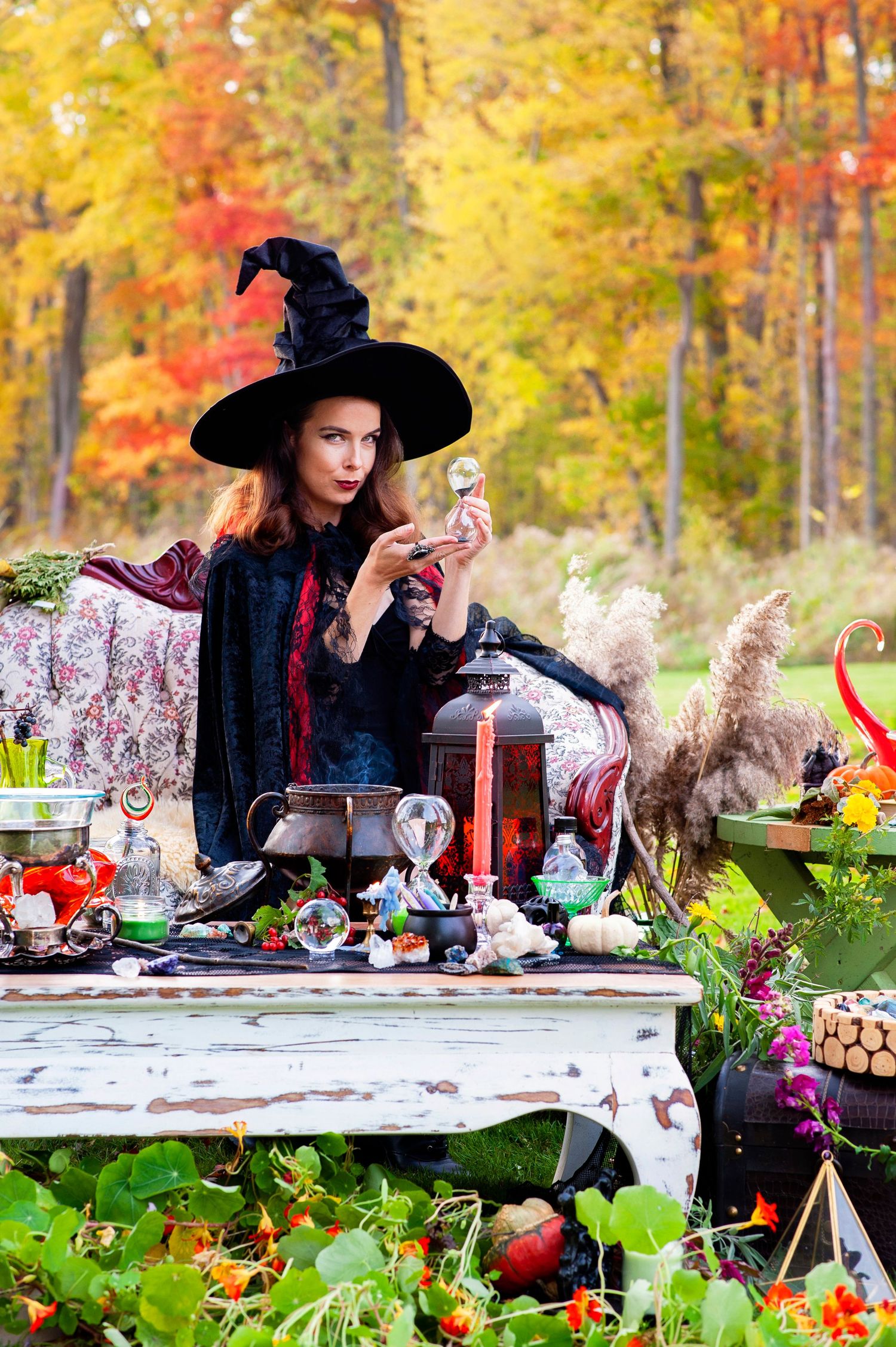 Woman dressed as a witch in black hat and cape stands at an outdoor table with potions and cauldron in autumn forest.