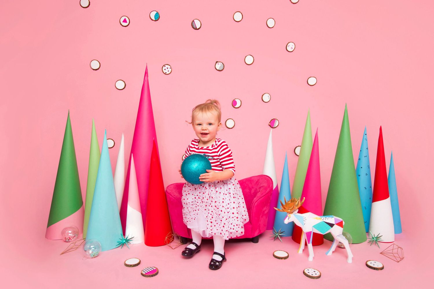 Cute toddler girl sitting among colorful paper Christmas trees on a bright pink holiday photo backdrop.