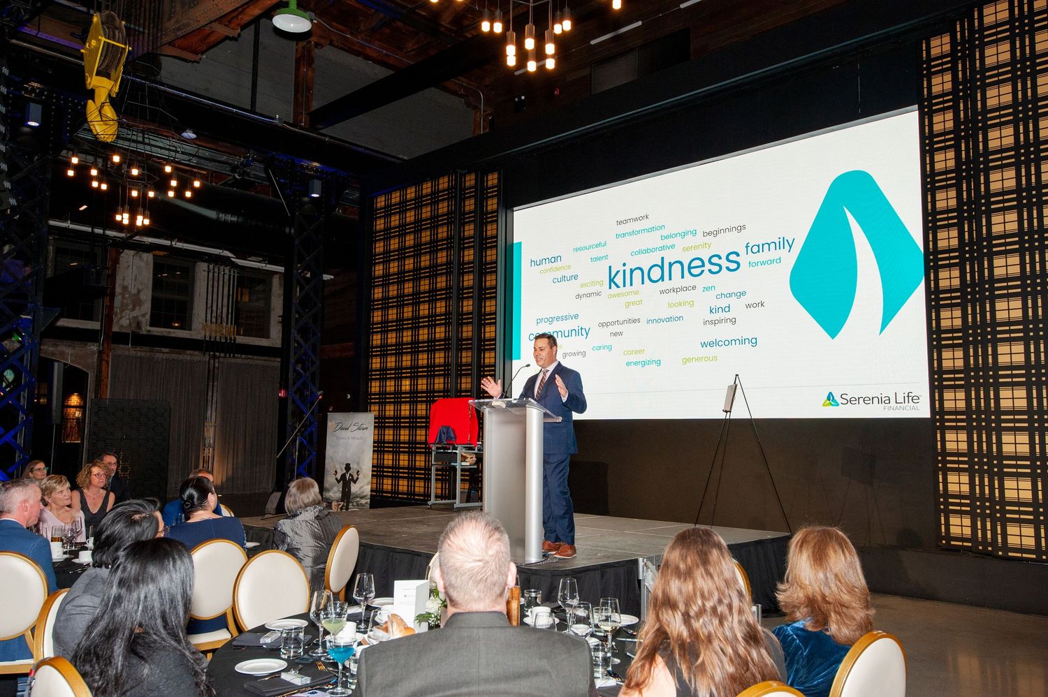 Speaker presents at podium before a screen displaying a kindness-themed word cloud at gala.