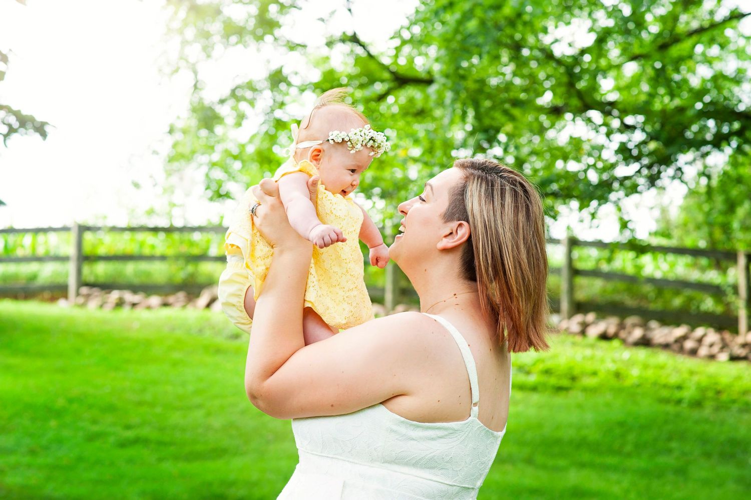 Young mother lifting smiling baby girl in yellow dress outdoors in sunny green garden.