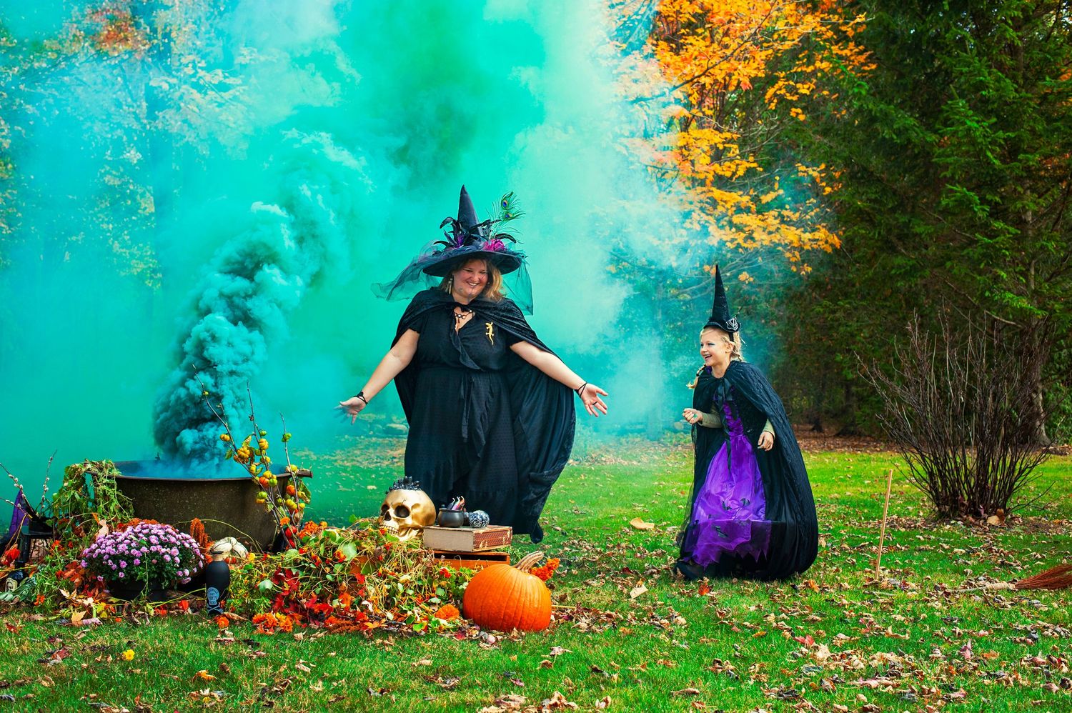 Two people dressed as witches with green smoke in autumn outdoor setting.
