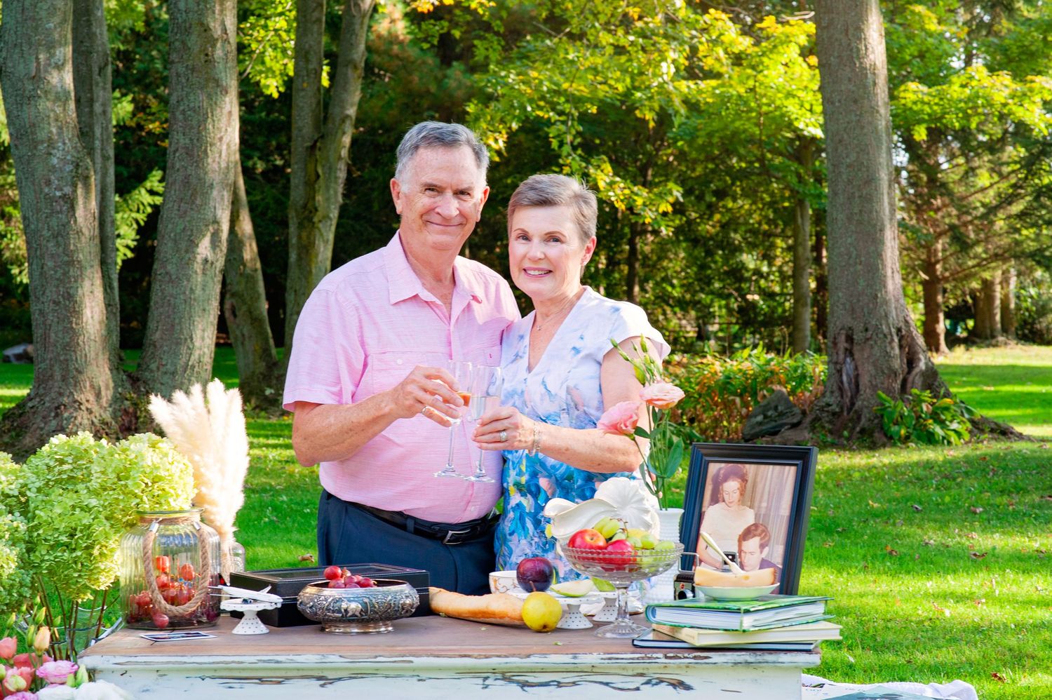 Happy senior couple toasting with champagne at outdoor table with fruits and flowers.