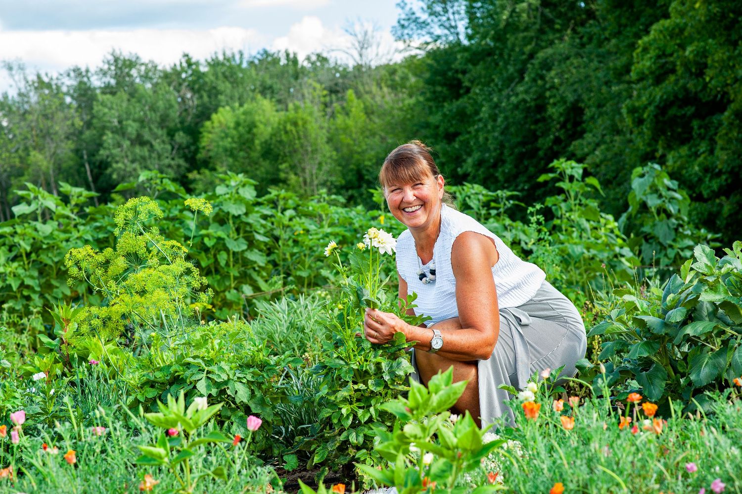 Smiling woman crouching in a lush green garden holding freshly picked flowers on a sunny day.