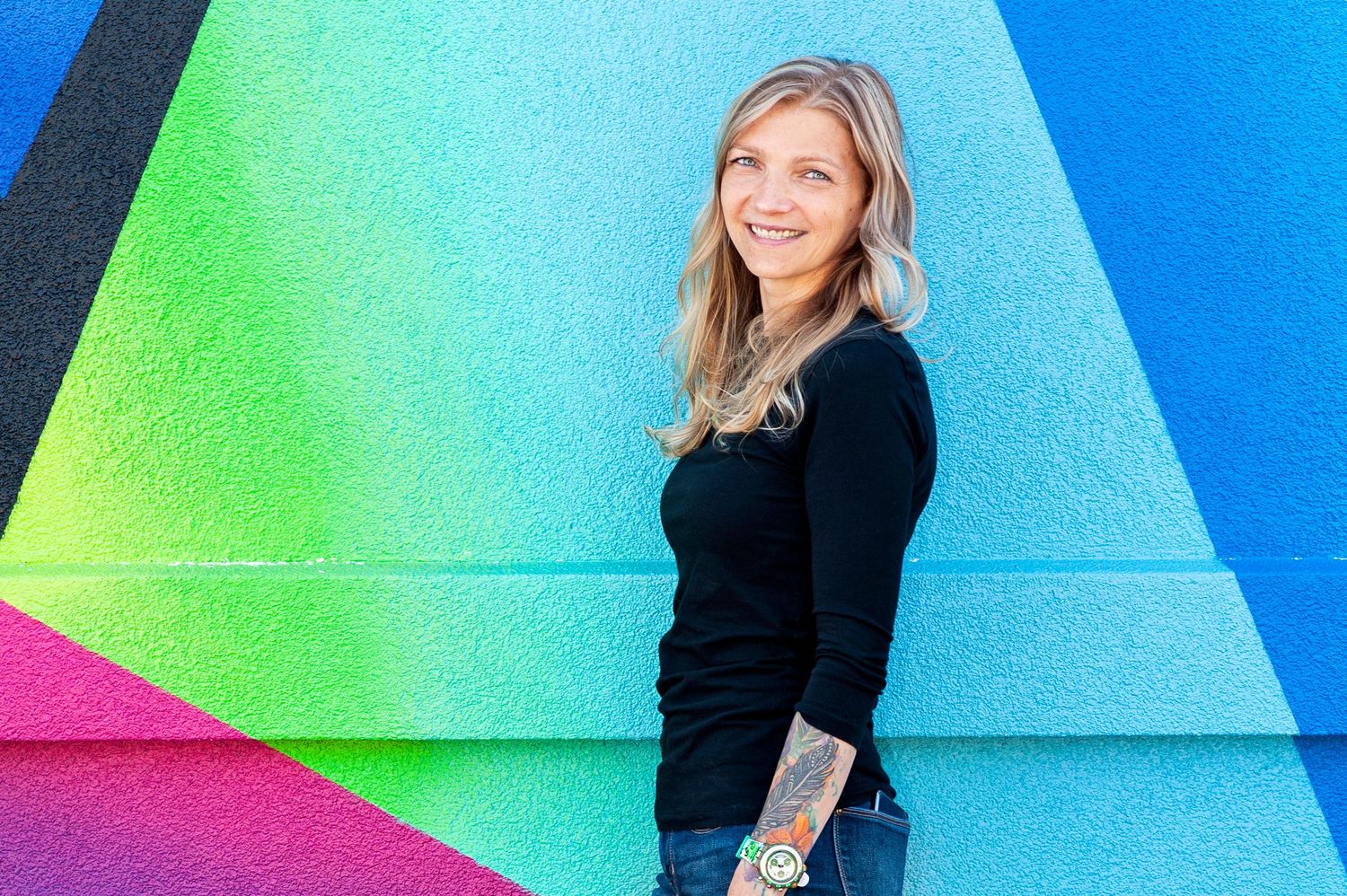Smiling blonde woman in black top standing against colorful geometric mural wall.