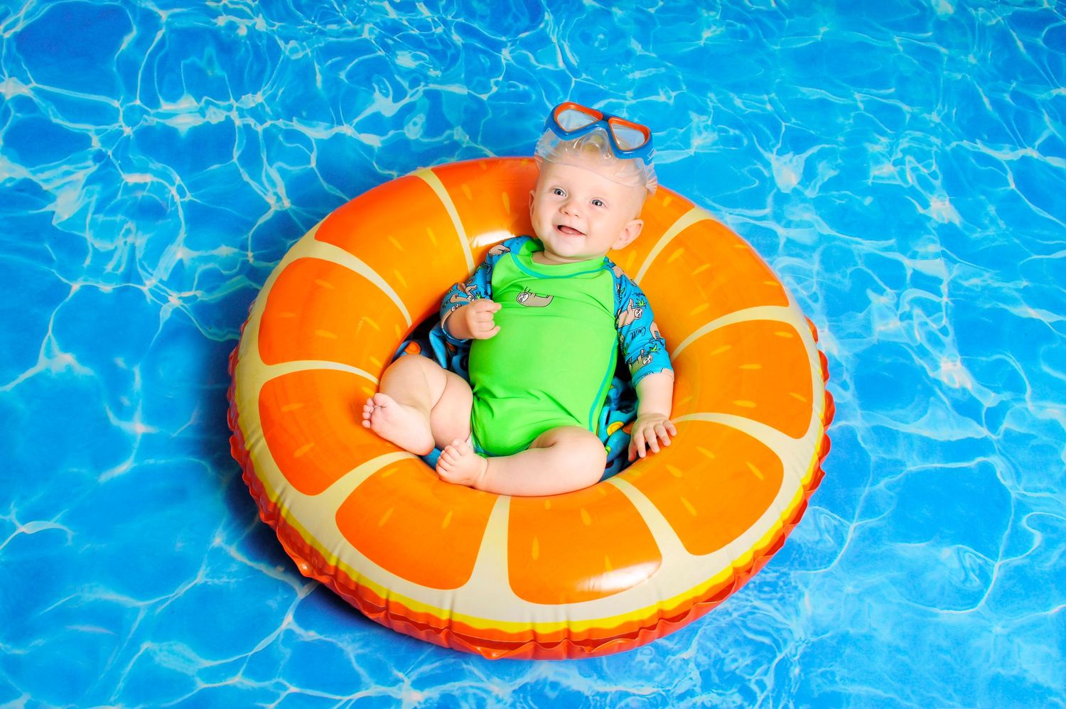 Cute baby in green swimsuit sitting in an orange inflatable ring float in a blue pool.