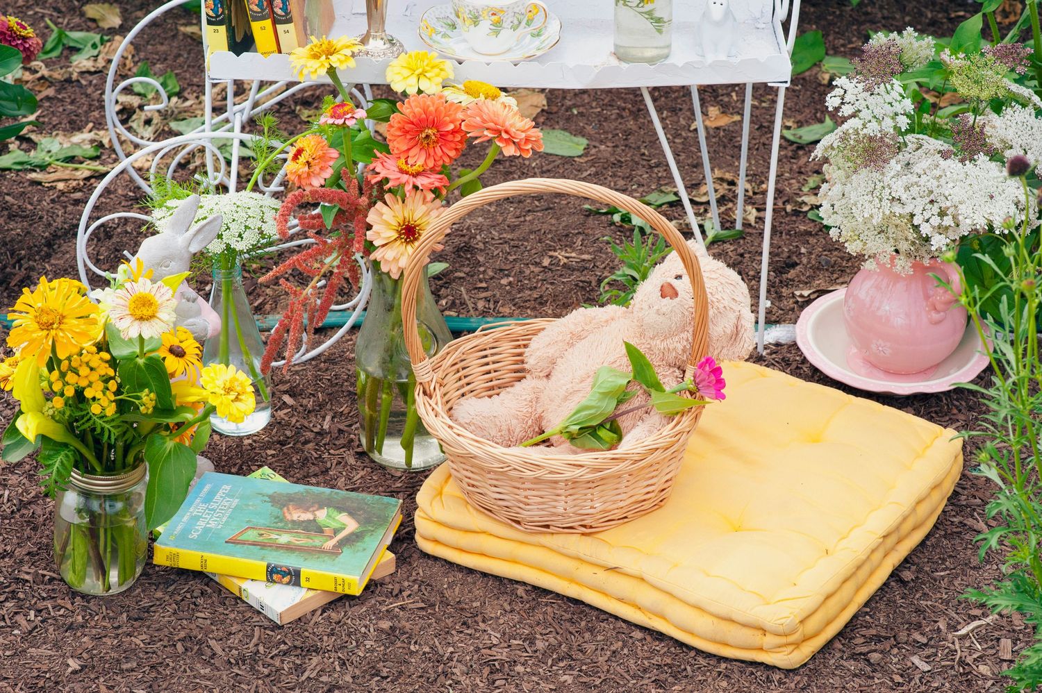 Wicker basket with teddy bear surrounded by colorful flowers, books, and garden decor on soil.