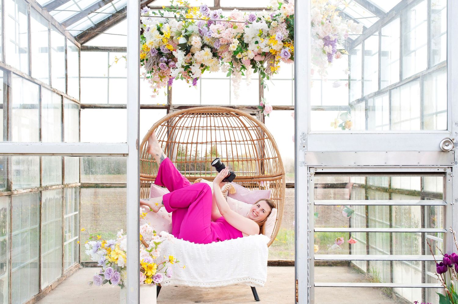 Woman in pink pants relaxes in hanging rattan egg chair inside a flower-decorated greenhouse.