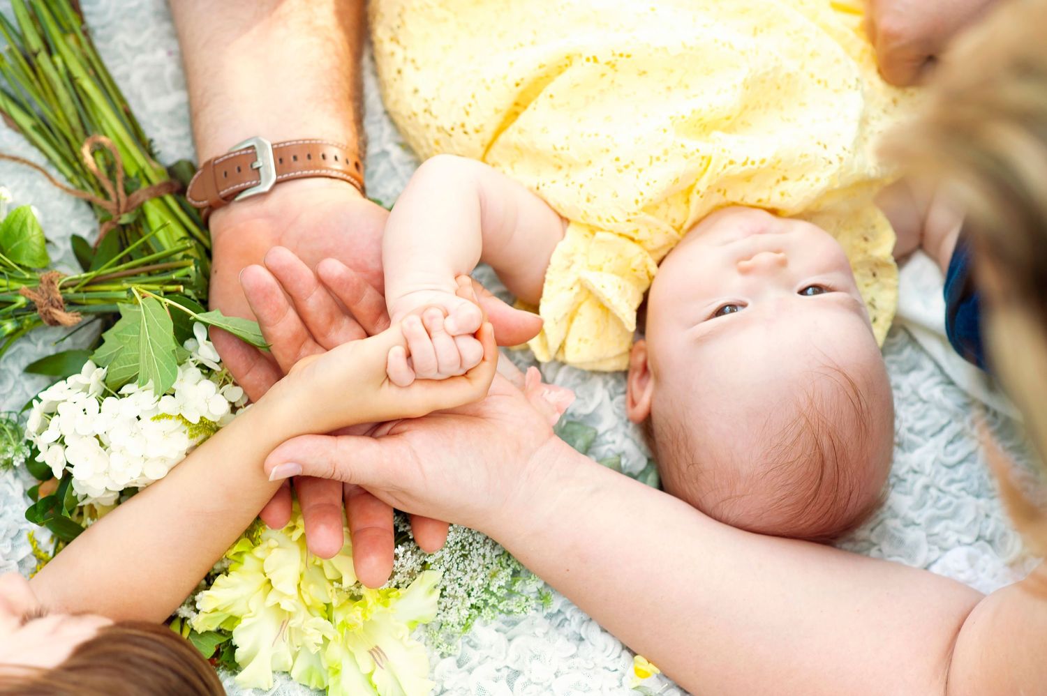 Family hands gently holding newborn baby in yellow outfit surrounded by white flowers.