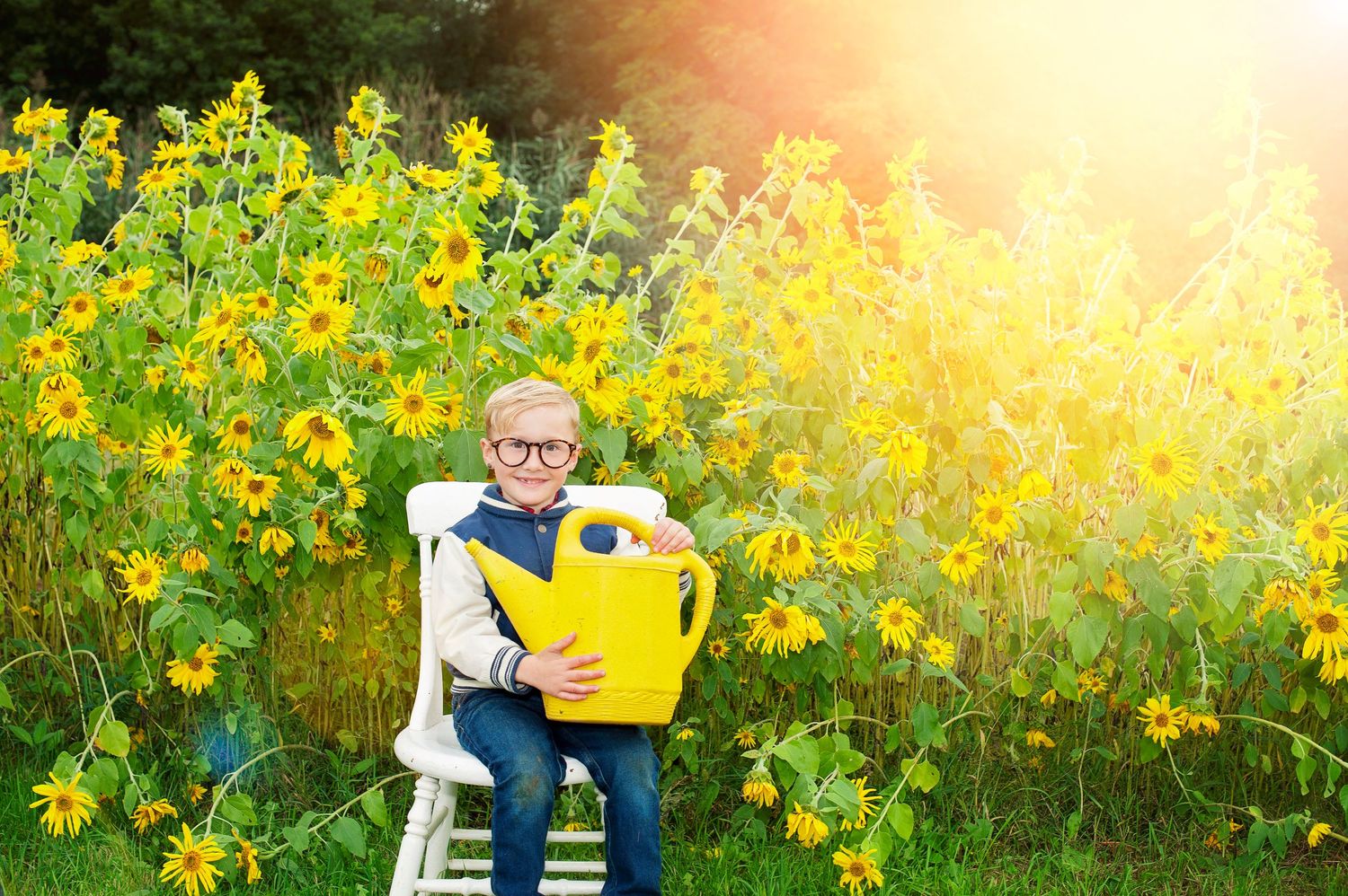 Young boy with glasses holding yellow watering can sitting in front of sunflower field in bright sunlight.