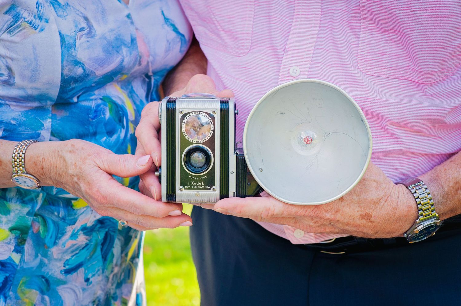 Couple holding vintage Kodak Duaflex camera outdoors in sunny garden setting.