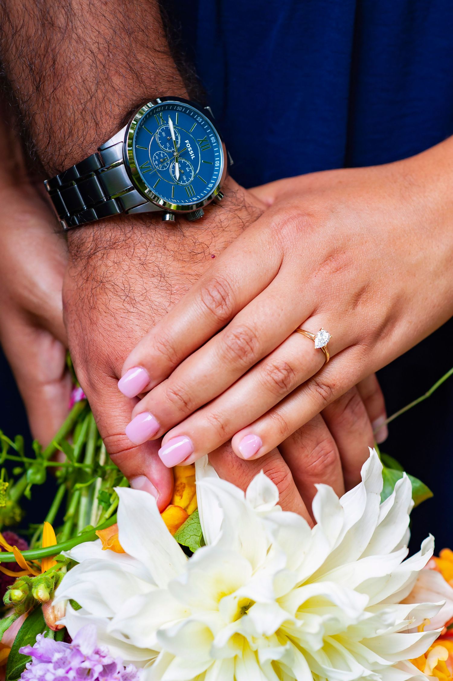 Close-up of engaged couple hands showing diamond ring and blue watch over flowers.