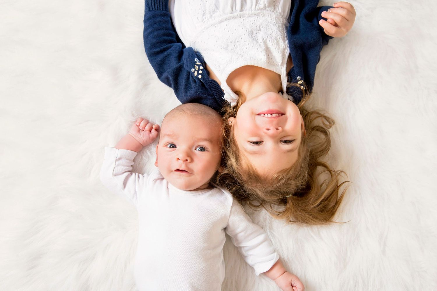 Newborn baby and young blonde child lying together on a white fluffy rug smiling upward.