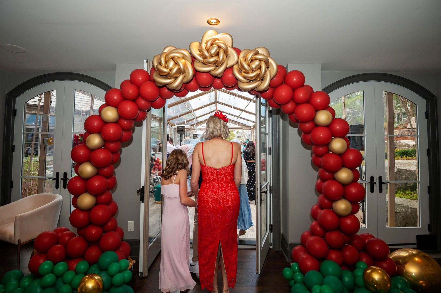 Woman in red dress and child walking through an elegant red and gold balloon rose arch at a party entrance.