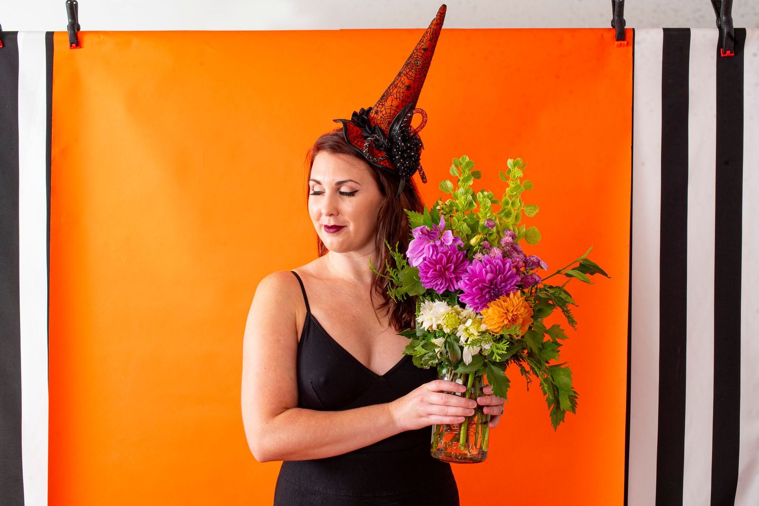 Woman in black dress and witch hat holding colorful flower bouquet against orange backdrop.