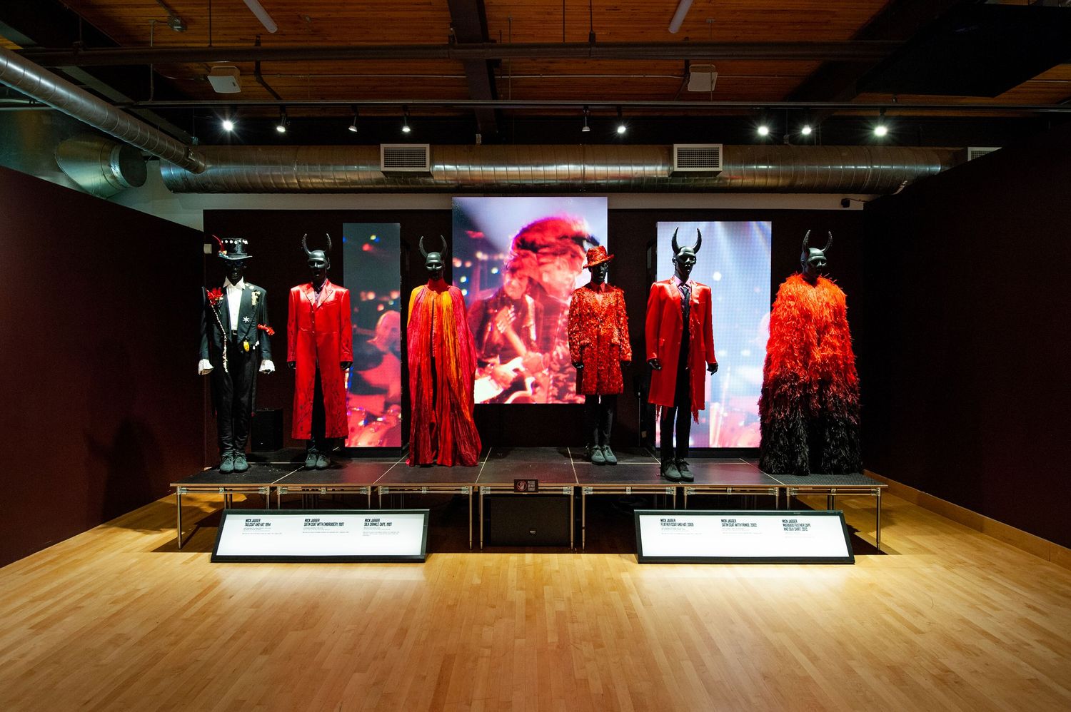 Red Rolling Stones stage costumes displayed in a museum exhibition with dramatic lighting and video screens.