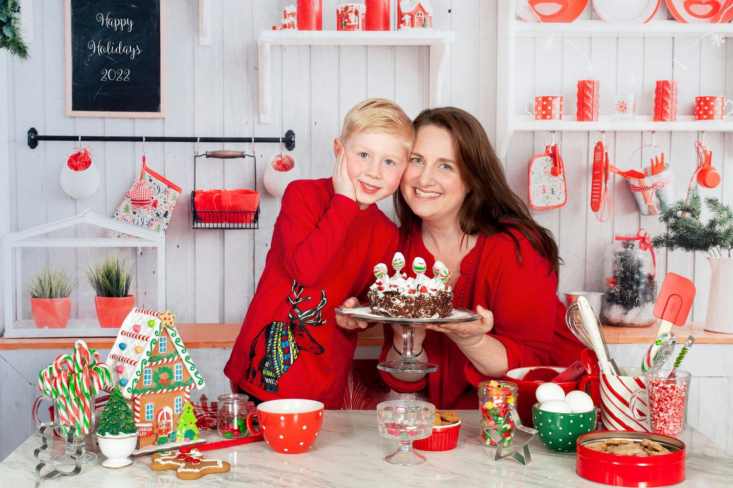 Mother and son in red outfits holding Christmas cake in festive holiday kitchen setting.