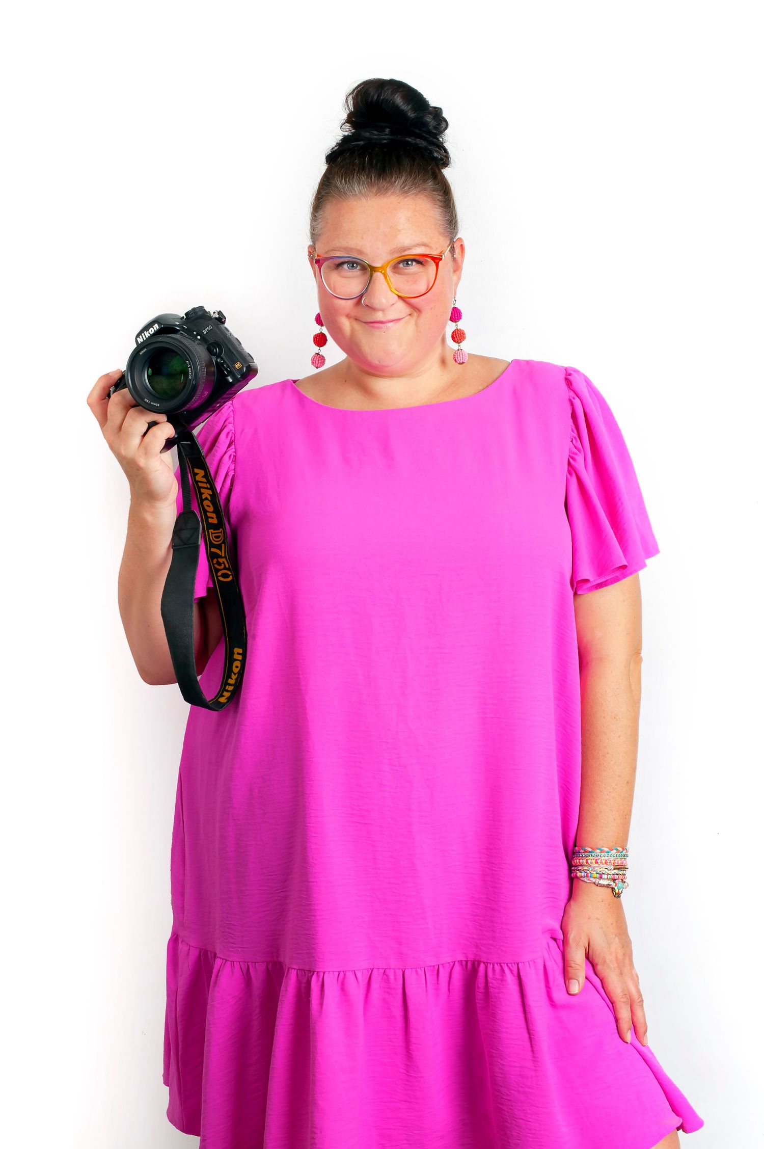 Woman in pink dress holding Nikon camera, wearing orange glasses and red earrings, smiling against white background.