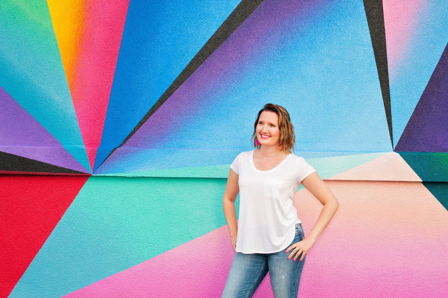 Smiling brunette woman in a white t-shirt and jeans posing against a vibrant colorful geometric mural.
