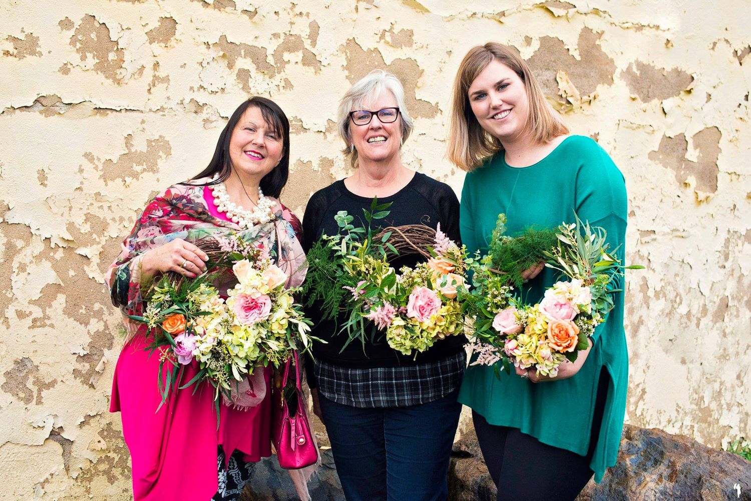 Three smiling women holding floral bouquets while standing against a rustic peeling paint wall.