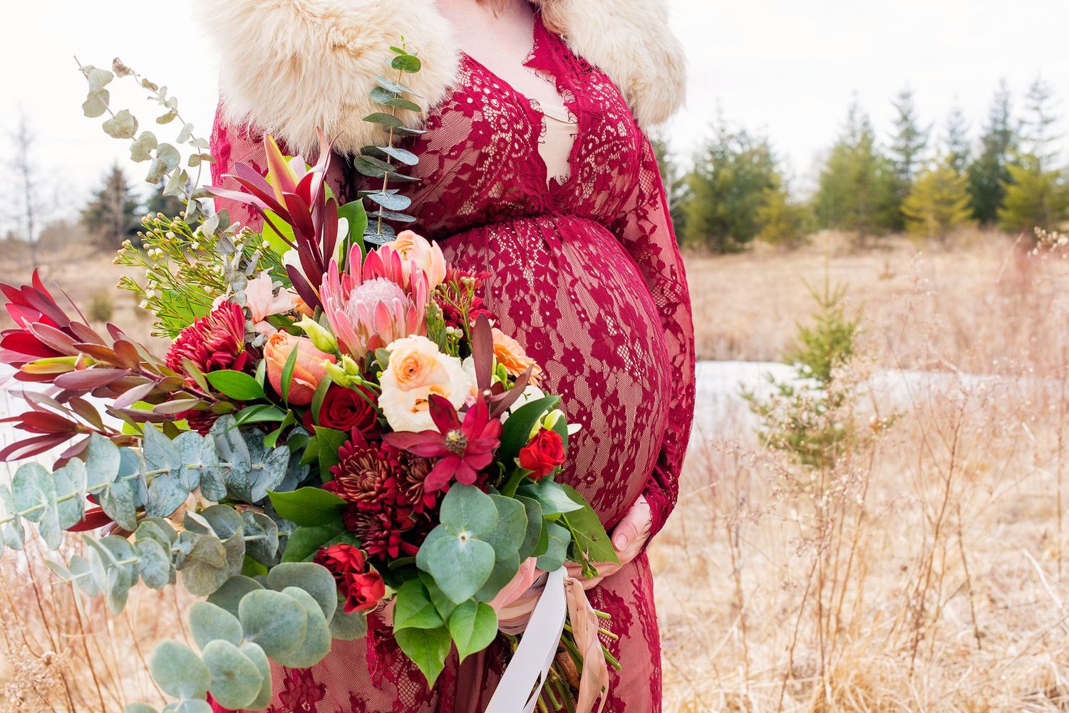Pregnant woman in red lace dress holding a lush bouquet of protea and eucalyptus outdoors in winter.