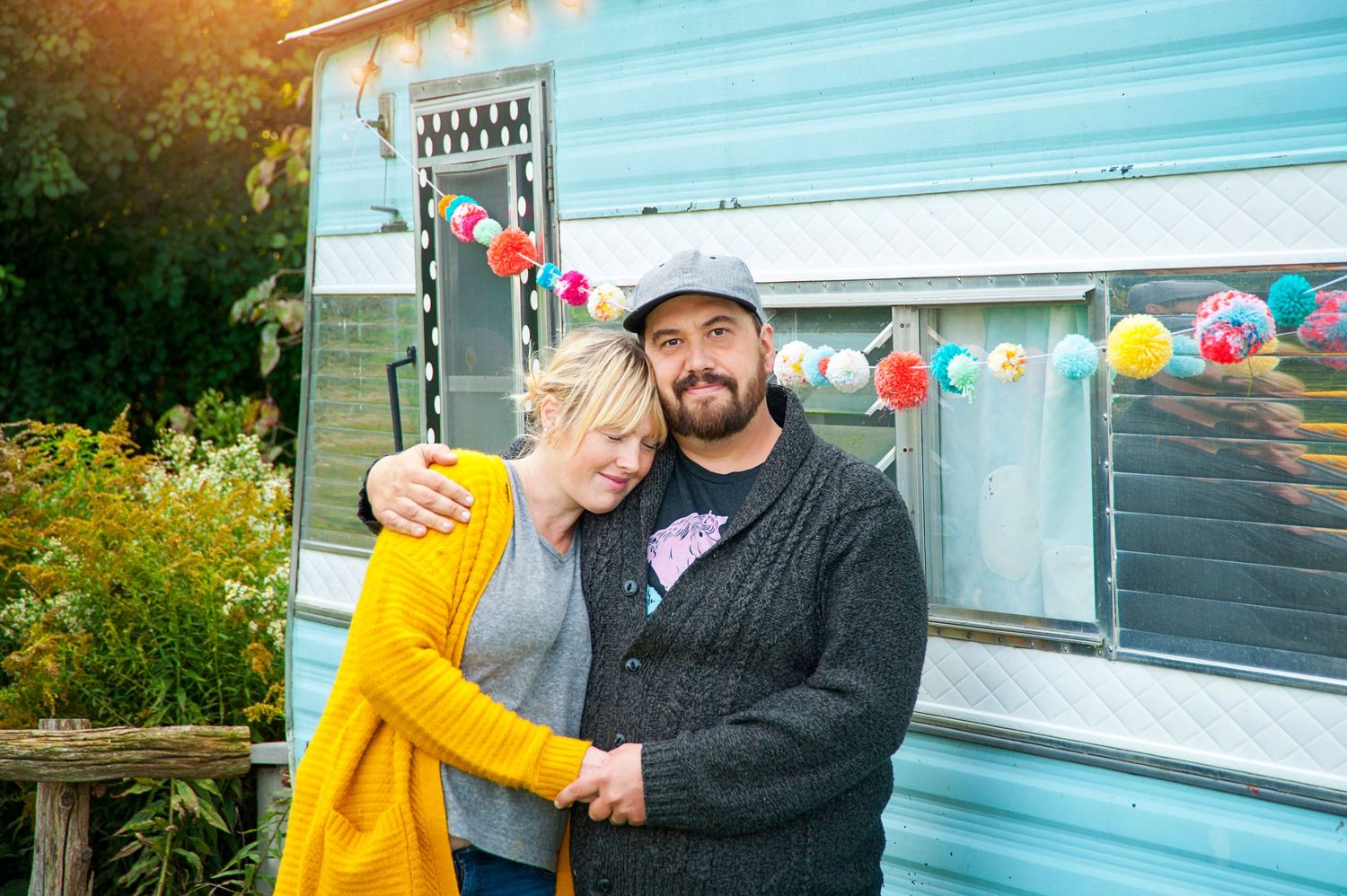 Couple embracing in front of light blue caravan decorated with colorful bunting garland.