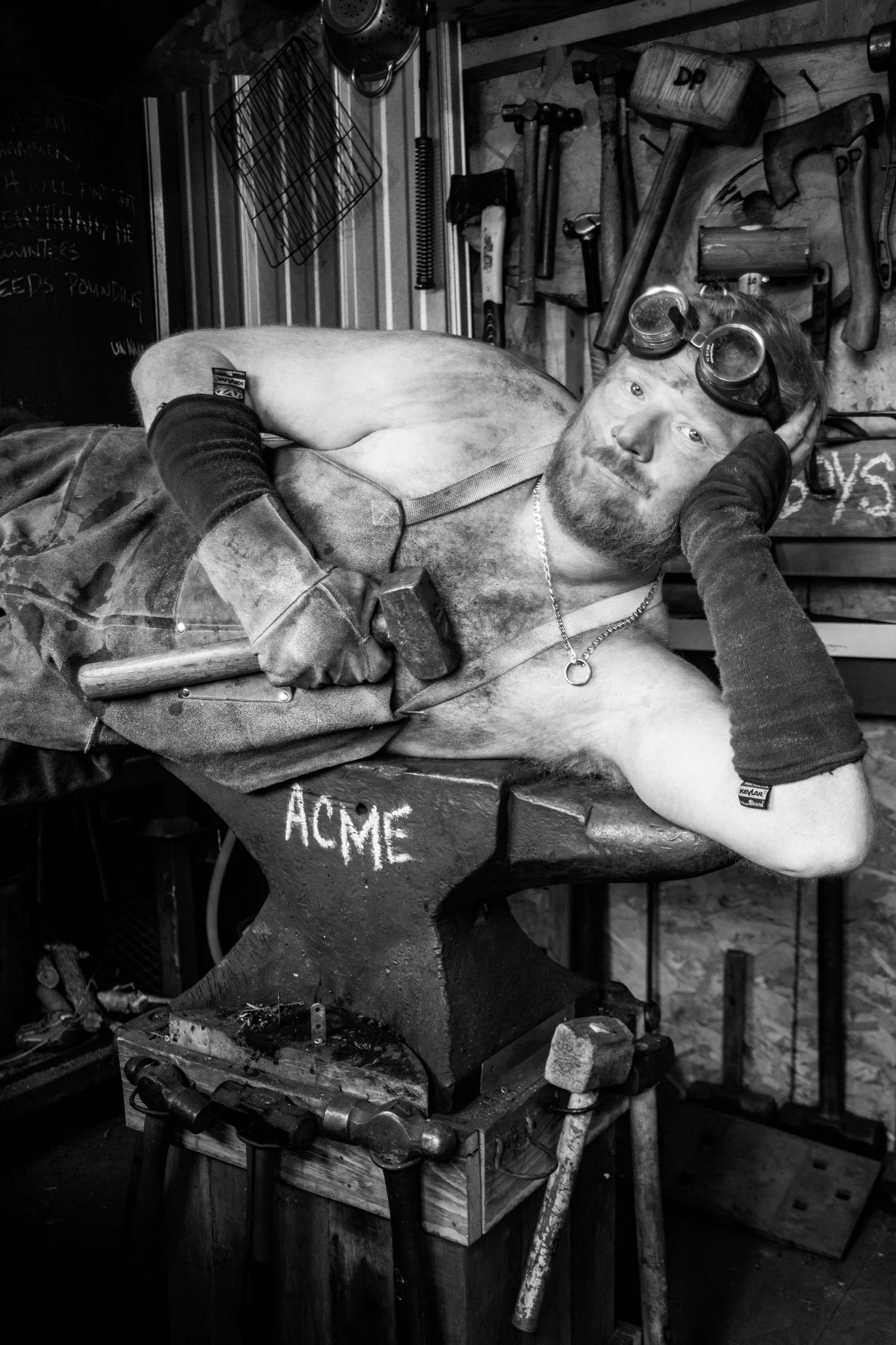 Shirtless bearded man lying across an ACME anvil surrounded by tools in a workshop, black and white.