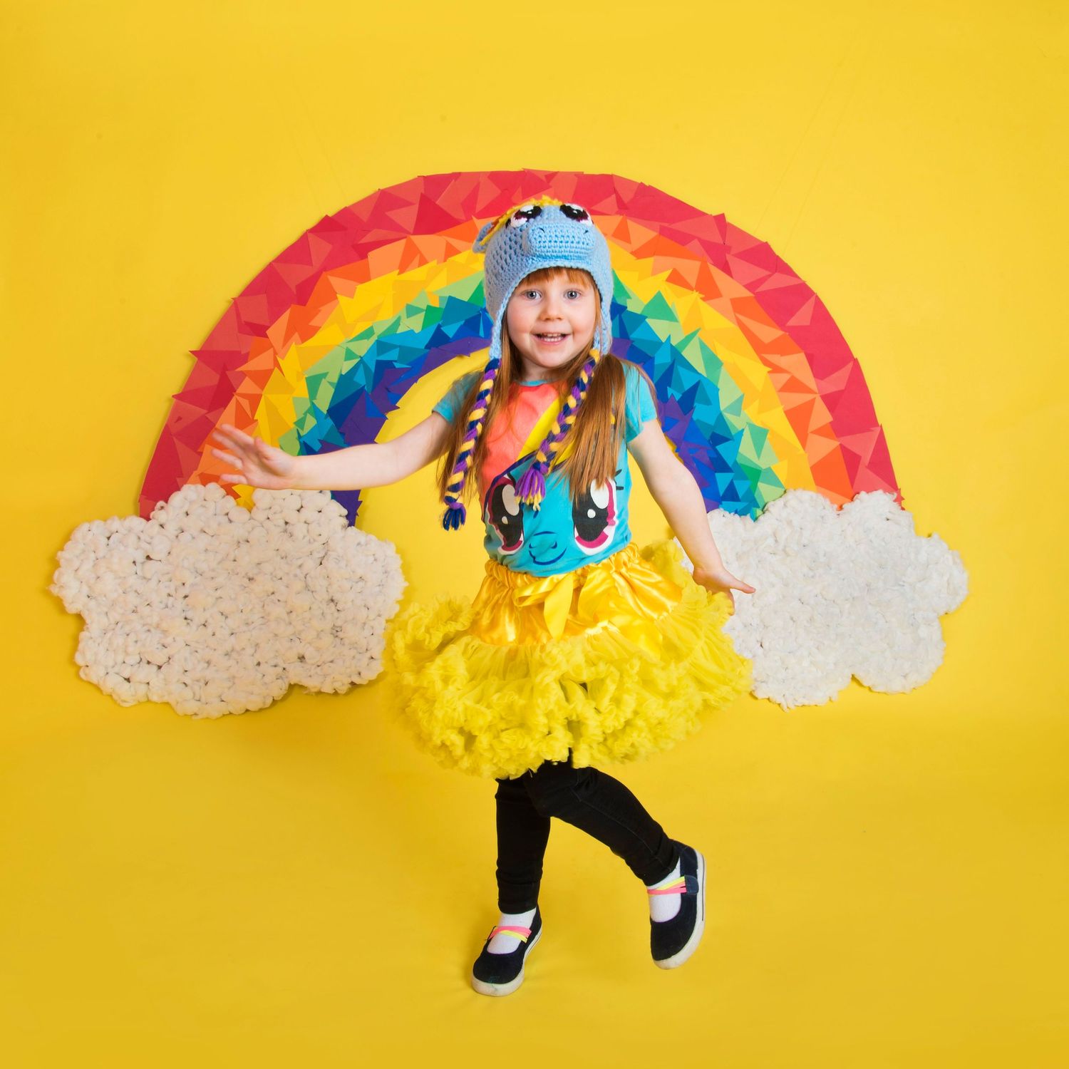Young girl in rainbow costume with yellow tutu, colorful hat, posing in front of a paper rainbow on yellow background.