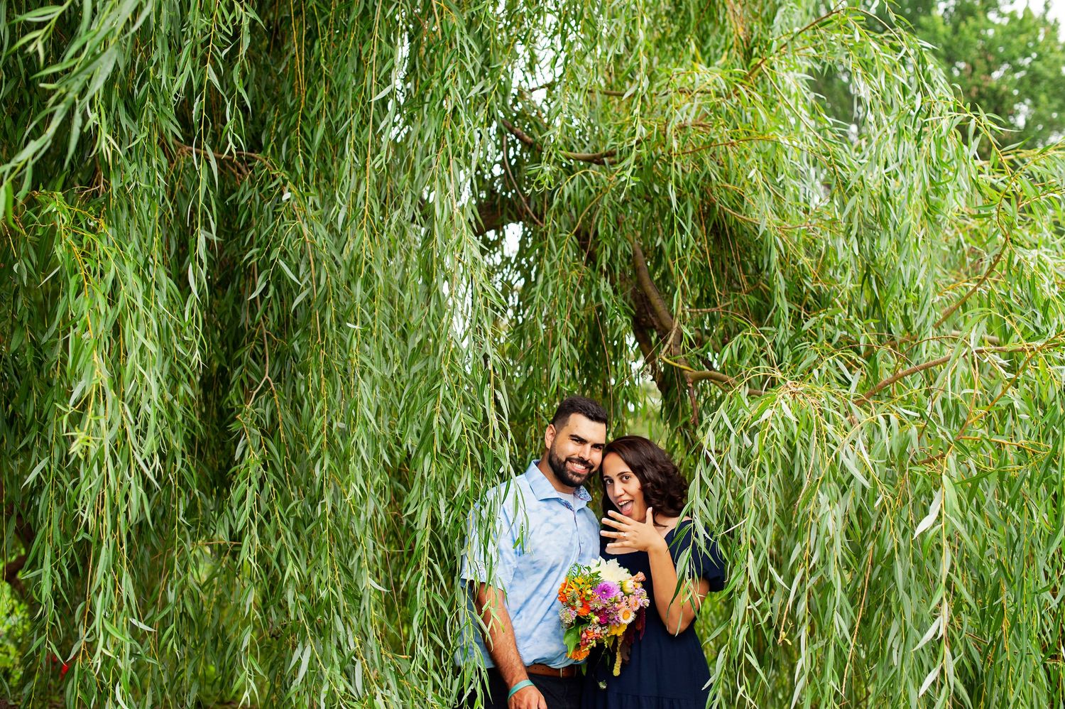 Romantic couple embracing under cascading weeping willow branches holding a bouquet.
