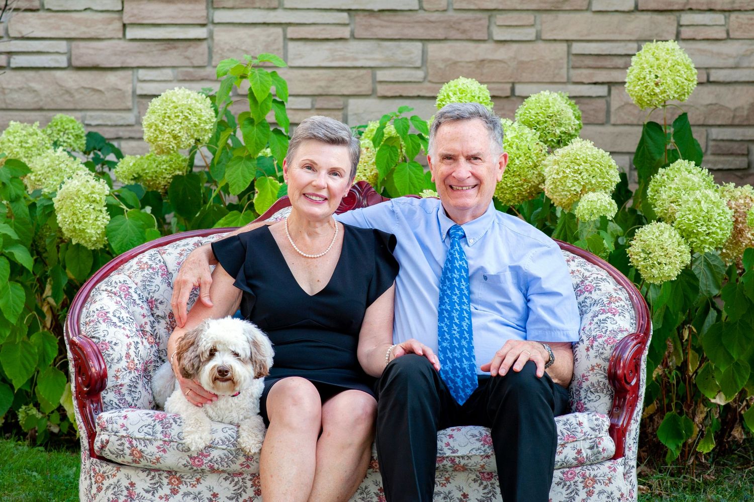 Senior couple sitting on floral sofa with small dog in front of hydrangea bushes.