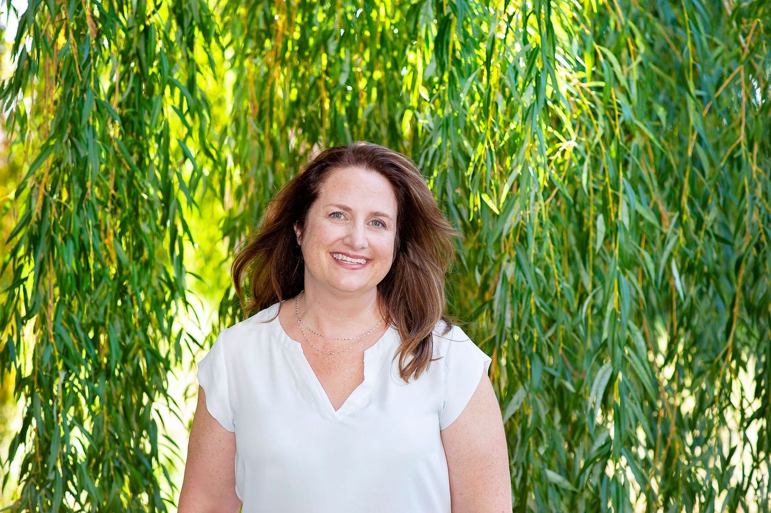 Woman smiling outdoors surrounded by lush green weeping willow branches in natural light.