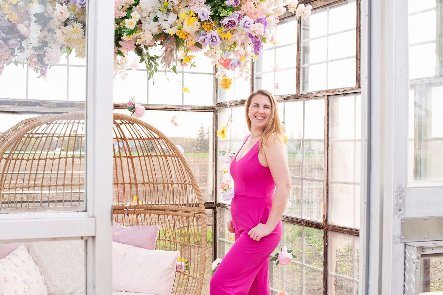 Blonde woman in pink dress poses near wicker egg chair in sunlit greenhouse with hanging floral arrangements.
