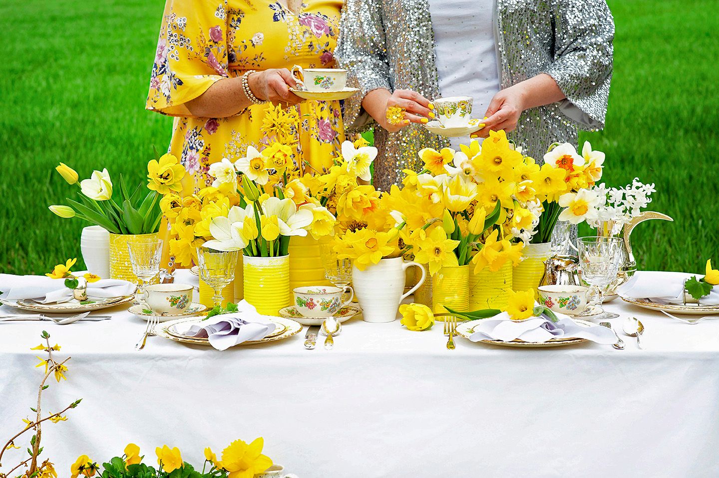 Two women in yellow floral outfits enjoying tea at an elegant outdoor spring table decorated with yellow daffodils.