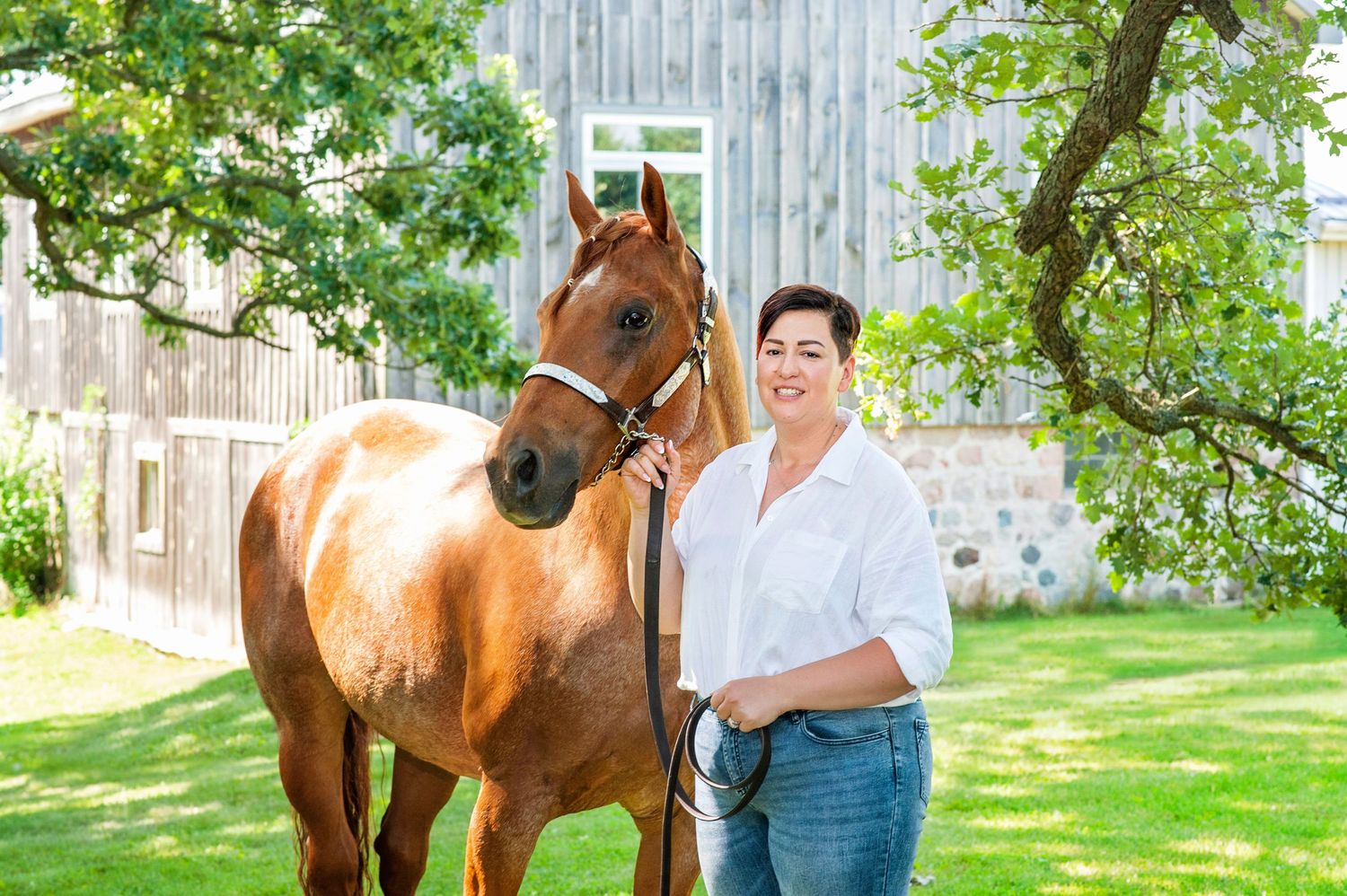 Smiling woman in white shirt standing beside a chestnut horse near a rustic barn.